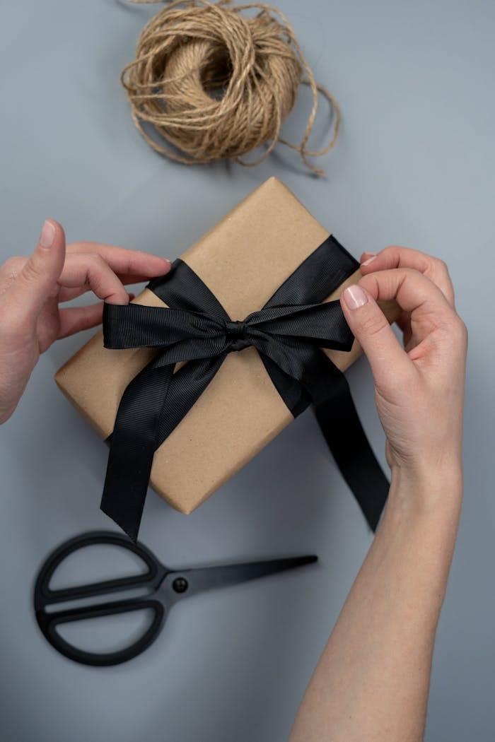 Services Hands tying a black ribbon on a craft paper gift box, next to scissors and twine.